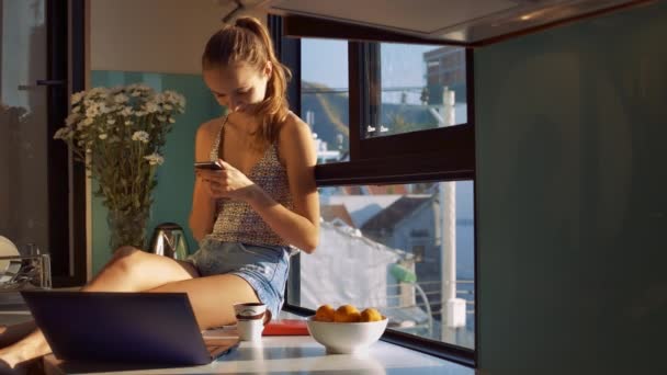 美丽的女孩微笑 并阅读新的信息在手机上的电脑和水果在窗台上早上 — 图库视频影像