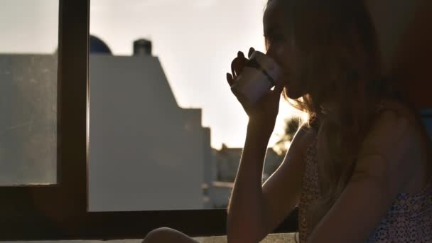 紧密的女人剪影与松散的流动的头发坐在窗台上喝热咖啡从杯反对明亮的日出 — 图库视频影像