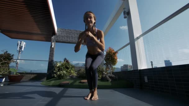 运动的年轻妇女与辫子改变现代屋顶休息室的瑜伽姿势对蓝天的人工植物 — 图库视频影像