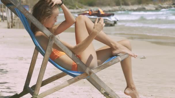 圧延の海の波とモーター ボートの砂浜に木製の折りたたみ椅子のクローズ アップ スリムなブロンドの女の子の日焼け — ストック動画
