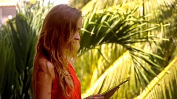 クローズ アップ側ビュー幸せな素敵な女の子が夏の日の太陽の下でヤシの木に対してタブレットを使用してインターネットをサーフィンします — ストック動画