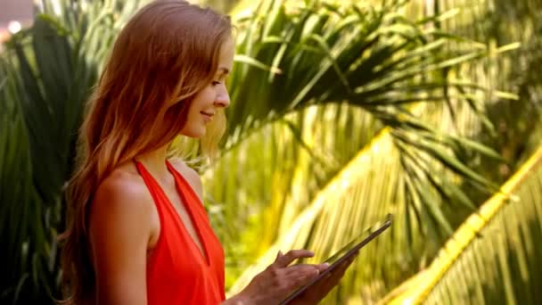 关闭侧面视图尼斯微笑长发女孩冲浪互联网与片剂附近棕榈叶下明亮的阳光下 — 图库视频影像