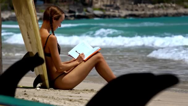 クローズ アップ サイドビュー少女海岸に座っているし 平和の海に対しての日記にメモを書いて幸せ — ストック動画