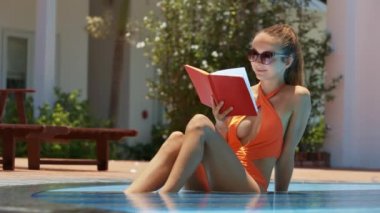Turuncu bikini kırmızı kırmızı kitap günlüğü resort otel sığ yüzme havuzunda oturan sarışın Bayan