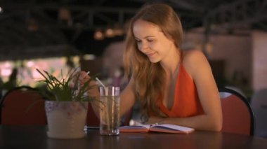 Kırmızı elbiseli portre sarışın uzun saçlı Bayan tropikal pot bitki ile masada oturur ve kafede kokteyl içer