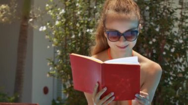 tatlı güzel sarışın Bayan modern güneş gözlüğü tropikal bush ile beyaz çiçekler karşı kırmızı kitap okur