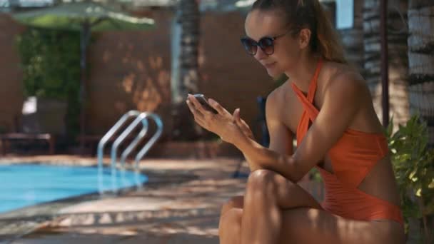 带马尾辫的金发女郎坐在游泳池附近阴凉的地方 短信在智能手机上 — 图库视频影像