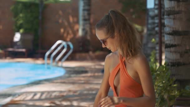 オレンジ ビキニでクローズ アップ スリムなブロンドの女の子は 晴れた日の日陰で座っている日焼け止めと腕をこすれ — ストック動画