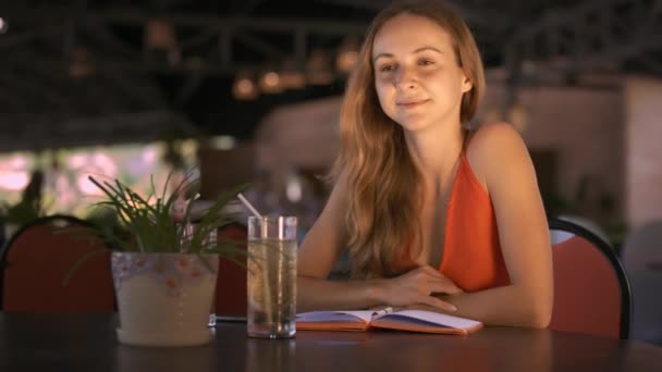 ポットの植物とジュースのメガネとカフェのテーブルに座っている緩い流れる髪のブロンドの長いスマート女の子ビューを閉じる — ストック動画