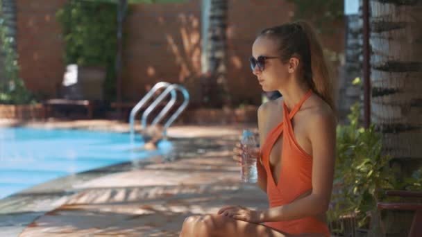 近观苗条女孩现代橙色比基尼在阳光明媚的日子里 从瓶子对酒店游泳池喝水 — 图库视频影像