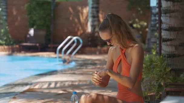 现代比基尼的年轻女子使用防晒霜涂抹手和手臂坐在阴凉的游泳池 — 图库视频影像
