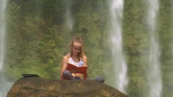 美丽的长发女孩看着红纸书的笔记本上的大老悬崖在国家公园反对瀑布 — 图库视频影像