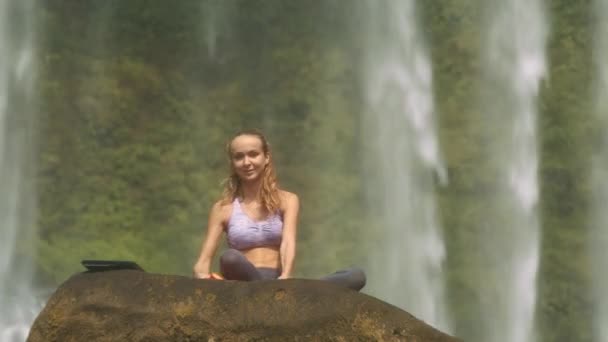 ブロンドの女の子は赤い本をわき置くし はドローンの滝の背後にある緑の植物に対して巨大な岩でリモート制御 — ストック動画