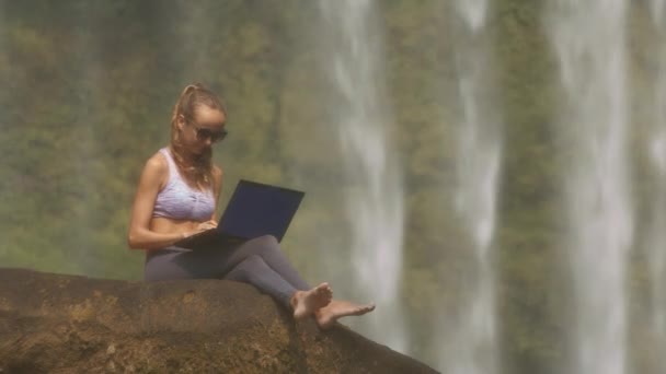 漂亮苗条的女孩在太阳镜和紫罗兰色顶部与笔记本电脑在大岩石反对著名瀑布 — 图库视频影像