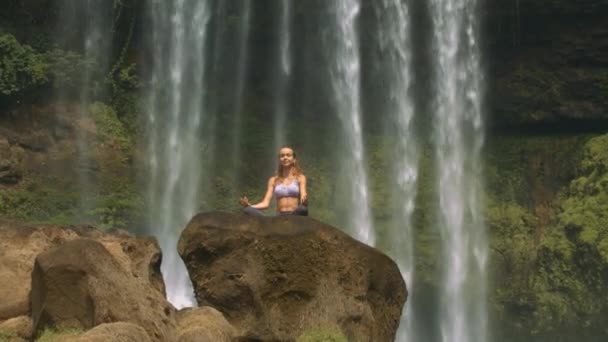 图片视图漂亮的金发长发女士把手臂放在膝盖上 放松瑜伽姿势上的褐色岩石对瀑布 — 图库视频影像