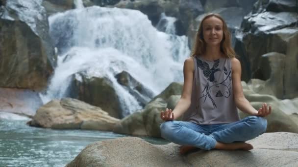 Блондинка в позе йоги на камне против водопада — стоковое видео