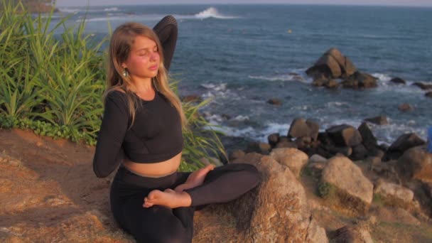 Lady in tracksuit sits on cliff in yoga pose against sea — 비디오