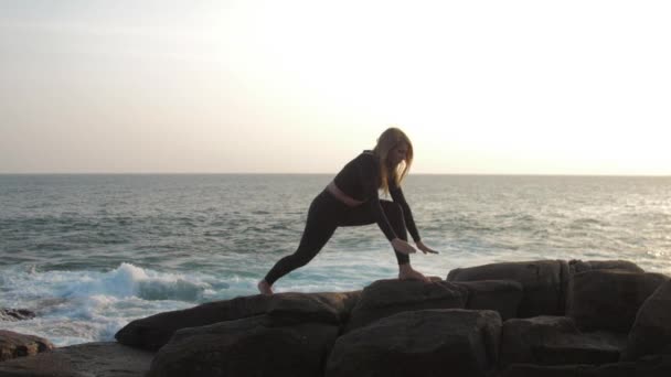 Young woman with long hair stretches on grey rocky cliff — 비디오