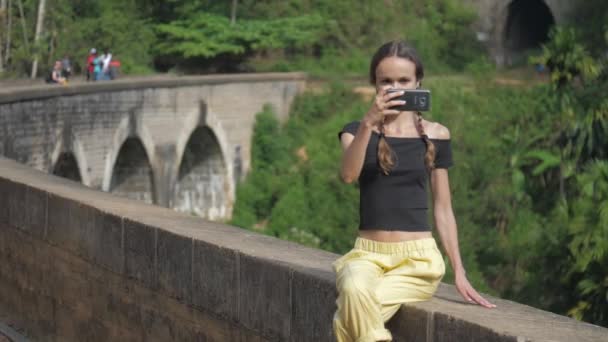 Chica en negro top hace selfie sentado en puente barrera — Vídeo de stock