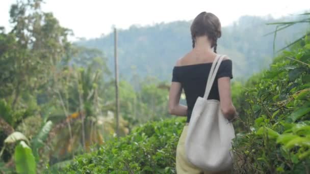 Молодая женщина с сумкой ходит по тропинке и пахнет чайным листом — стоковое видео