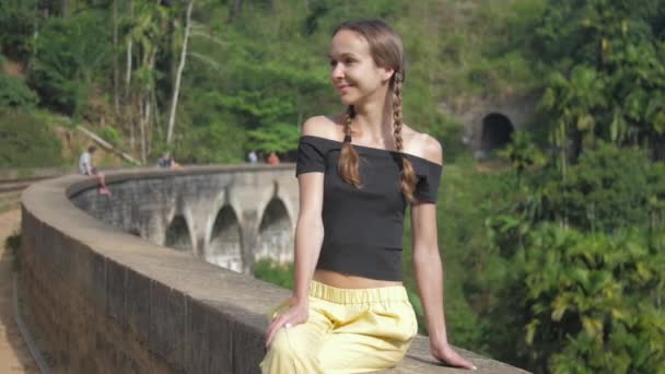 Menina senta-se na ponte histórica sorrindo contra a floresta selvagem — Vídeo de Stock