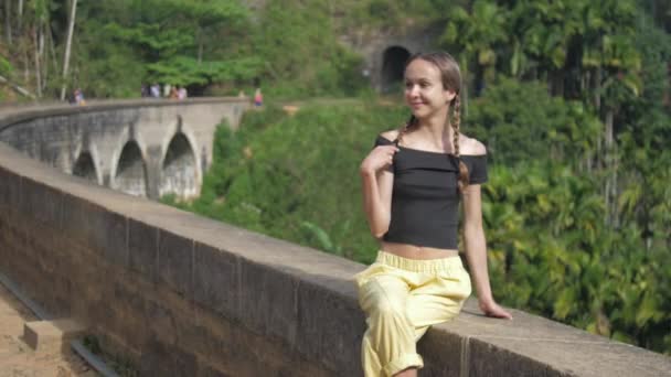 Meisje in zwart top zit op oude brug glimlachen tegen bos — Stockvideo