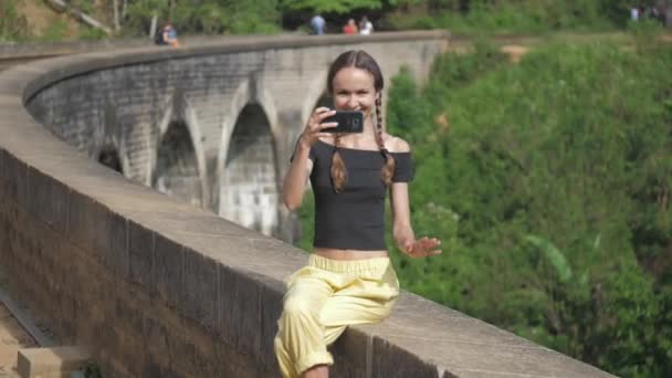 Girl makes selfie sitting on bridge against wild jungle — Stockvideo