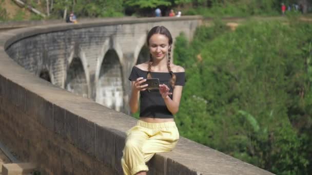 Girl makes selfie sitting on old bridge barrier by jungle — Stockvideo