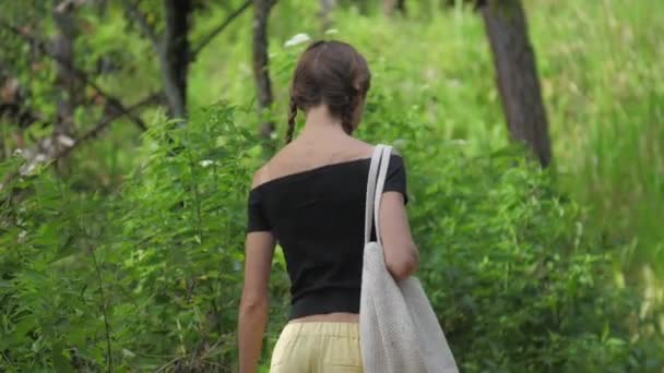 Young woman wanders along green forest holding white bag — Stock Video