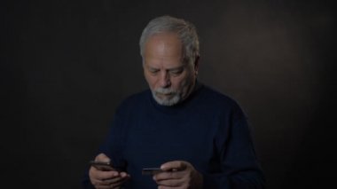 Yaşlı adam gri stüdyo geçmişi için kredi kartı ve akıllı telefon kullanıyor.
