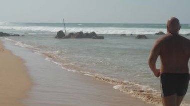 Avrupalı yetişkin bir adam güneşli bir günde okyanus kıyısında kumlu sahilde koşuyor.