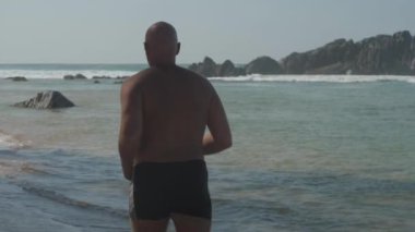 Avrupalı yetişkin bir adam güneşli bir günde okyanus kıyısında kumlu sahilde koşuyor.