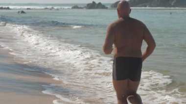 Avrupalı yetişkin bir adam güneşli bir günde okyanus kıyısında kumlu sahilde koşuyor.