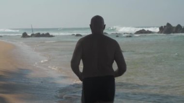 Avrupalı yetişkin bir adam güneşli bir günde okyanus kıyısında kumlu sahilde koşuyor.