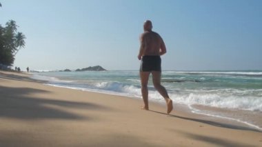 Avrupalı yetişkin bir adam güneşli bir günde okyanus kıyısında kumlu sahilde koşuyor.