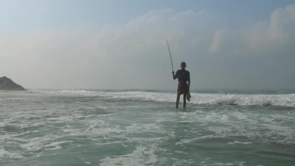 Rybář stojí v pěnící vodě oceánu pod zamračenou oblohou — Stock video