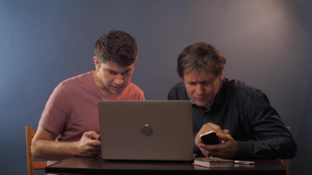 Молоді і зрілі чоловіки з телефонами дивляться на ноутбук на столі — стокове відео