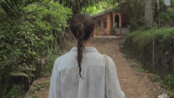 Κορίτσι περπατά κατά μήκος του δρόμου ανάμεσα σε τροπικά δέντρα προς την οικοδόμηση — Αρχείο Βίντεο
