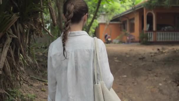 Девушка идет по тропе среди тропических деревьев к зданию — стоковое видео