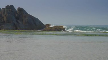 Keskin kahverengi kayaların yanında dalgalanan okyanus berrak mavi gökyüzüne karşı