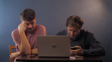 Babası olan adam masadaki bilgisayarın yanında akıllı telefon kullanıyor.