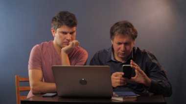 Oğlu dizüstü bilgisayarda çalışırken, babası akıllı telefonlara bakıyor.