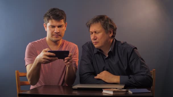 Homem maduro pergunta ao filho como usar o celular perto de uma mesa pequena — Vídeo de Stock