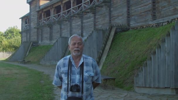Šedovlasý muž kráčí a mluví proti dřevěné pevnosti — Stock video