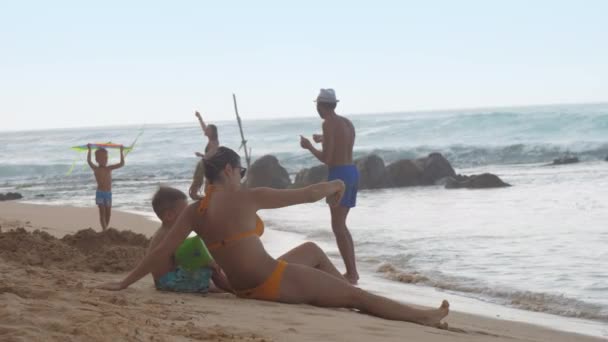 Dame se trouve sur la plage de sable près de garçon dans les flotteurs bras contre l'océan — Video