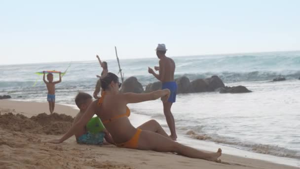 Lady sits on beach sand near boy in arm floats against ocean — Stock Video