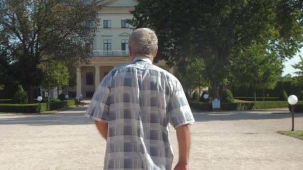 Homem de cabelos grisalhos caminha contra o palácio e exuberantes árvores verdes — Vídeo de Stock