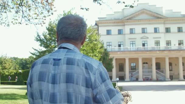 Šedovlasý muž kráčí proti paláci a svěží zelené stromy — Stock video