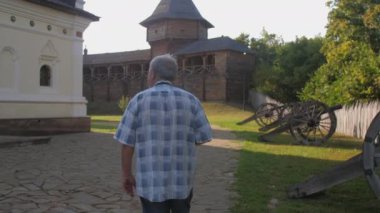 Emeklinin elinde siyah kamera var ve müze parkında fotoğraf çekiyor.