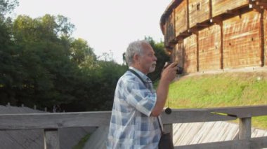 Emeklinin elinde siyah kamera var ve müze parkında fotoğraf çekiyor.